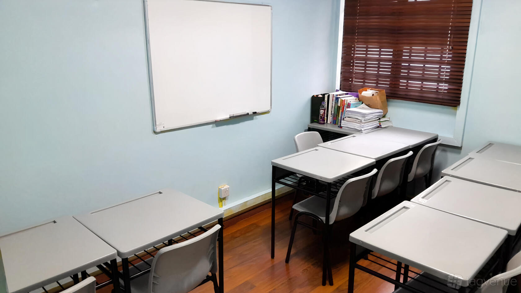 A classroom with gray desks, white plastic chairs, a wall-mounted whiteboard, and wooden flooring at Tampines West MRT Classroom Space.