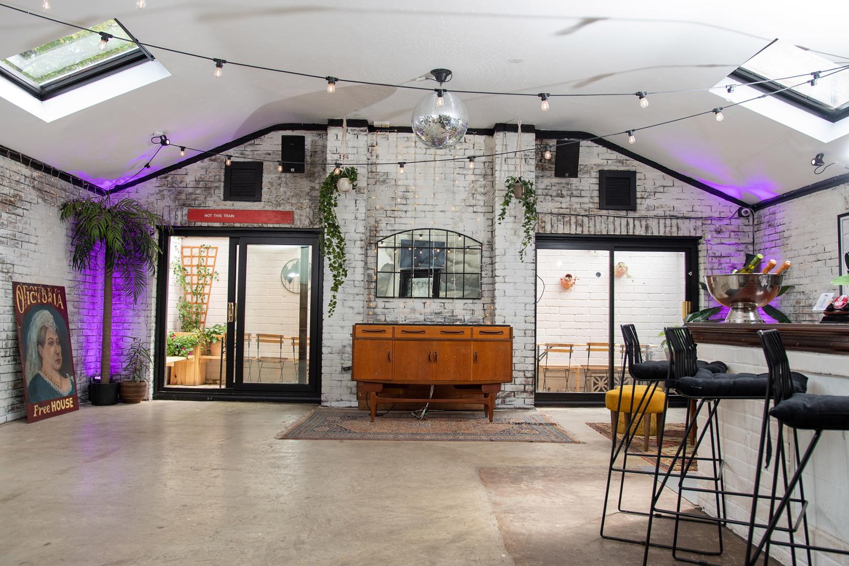 A function room in a pub with exposed white brick walls, skylights, bar seating, and purple accent lighting at The Clubhouse at The Vic.
