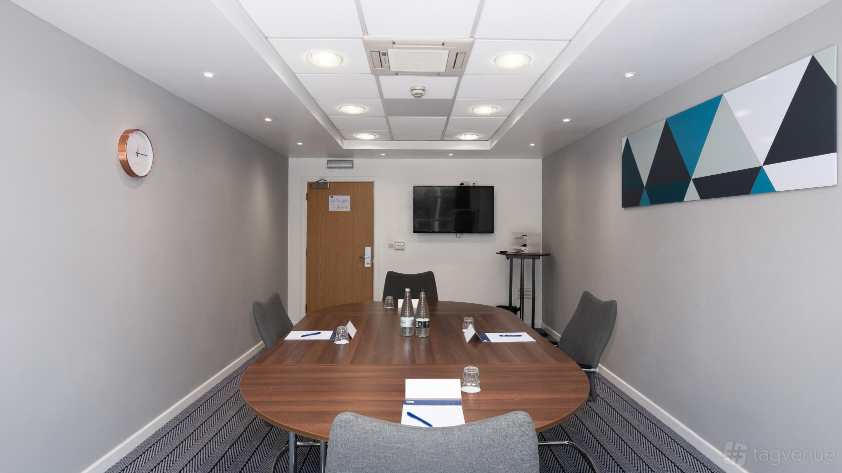 A meeting room with a large oval table, gray chairs, a wall-mounted TV, and geometric wall art at Holiday Inn Express Leigh Sports Village.