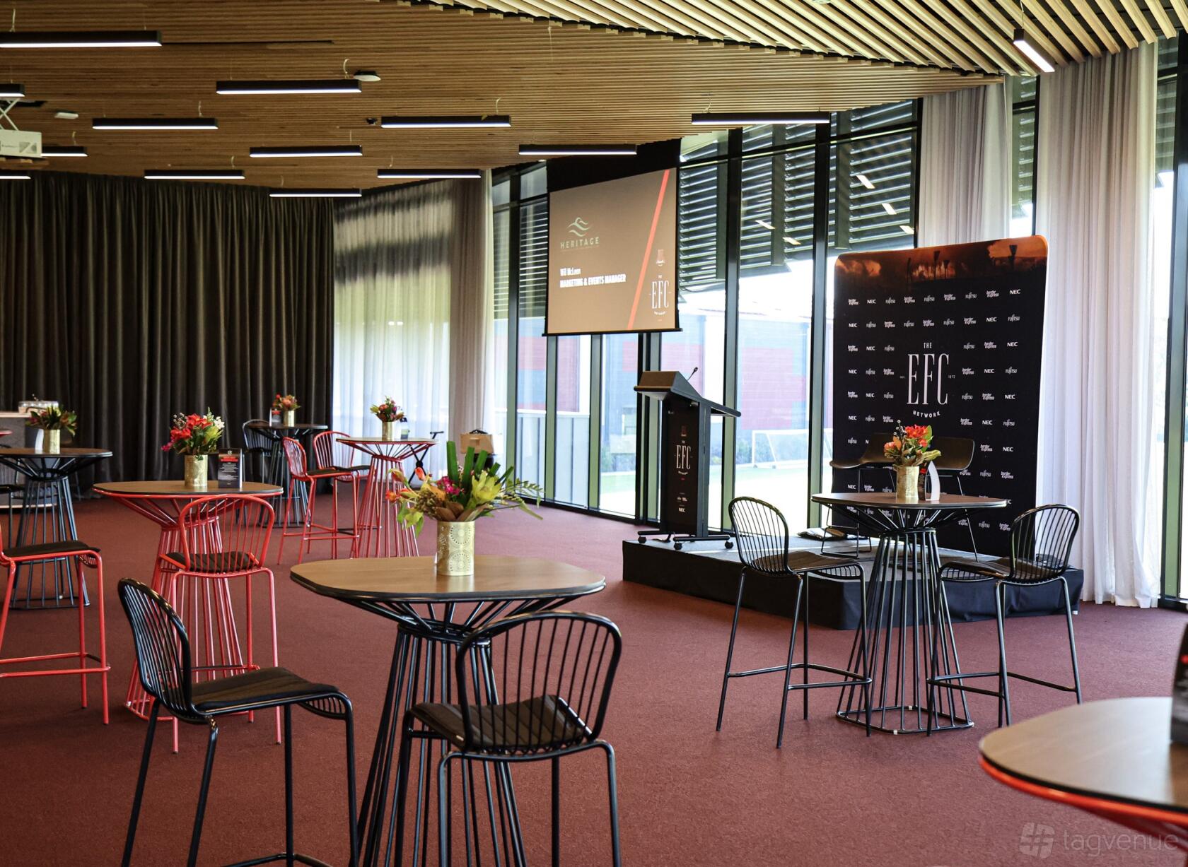 A function room with round cocktail tables, wire chairs, floor-to-ceiling windows, and a presentation stage at NEC Hangar - Essendon Football Club.