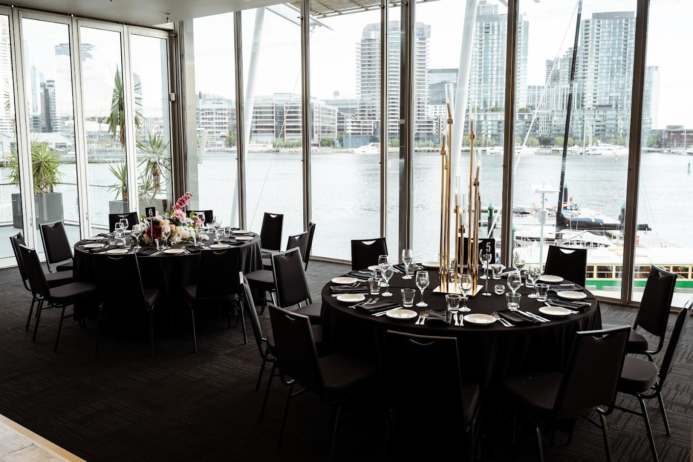 An event venue with floor-to-ceiling windows overlooking the waterfront and round tables set with glassware at The Promenade Docklands