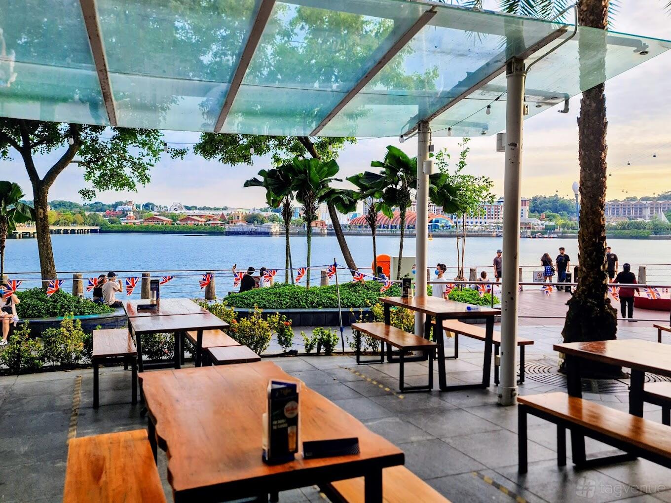 An open-air restaurant with wooden tables, views of the waterfront, and greenery at The Queen and Mangosteen.