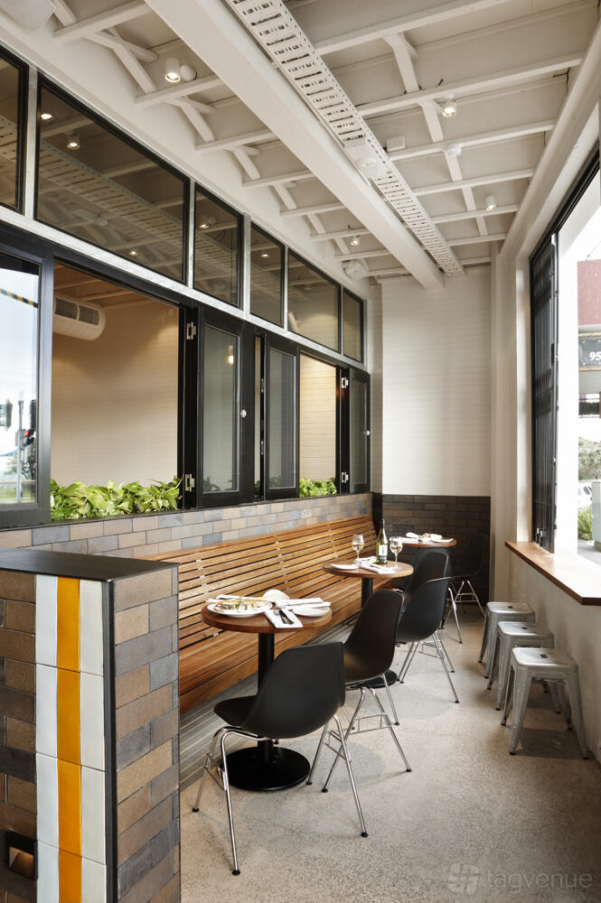 A restaurant terrace with wooden bench seating, small round tables set for dining, and a row of green plants at True South.