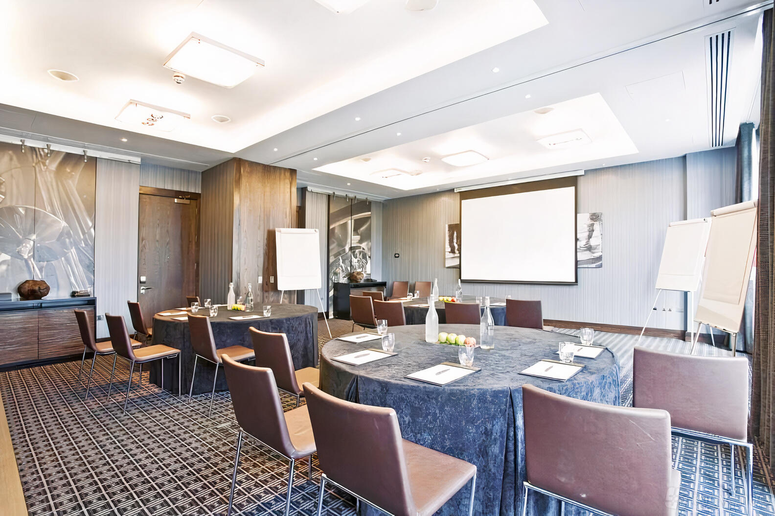A meeting room with round tables, brown chairs, flip charts, and a projector screen at Brooklands Hotel.