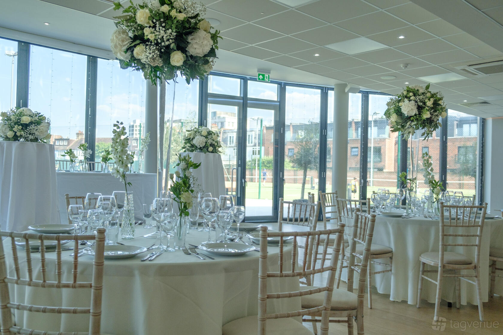 A function room with floor-to-ceiling windows, round tables with white linens, and floral centerpieces at Parsons Green Club.