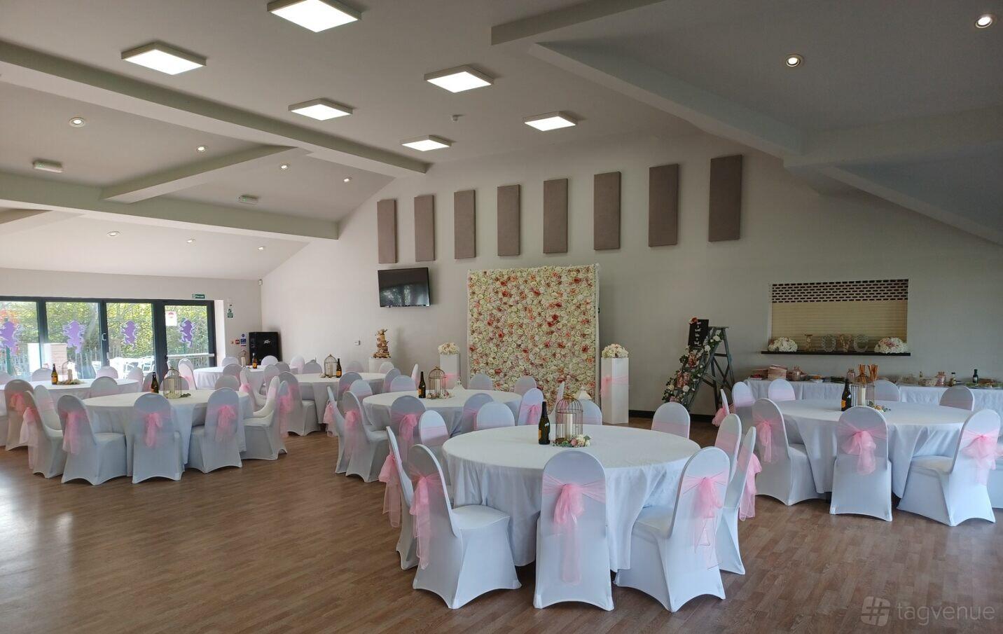A hall with round tables draped in white linens and pink chair sashes, wood flooring, and large windows at Leicester Lions RFC.
