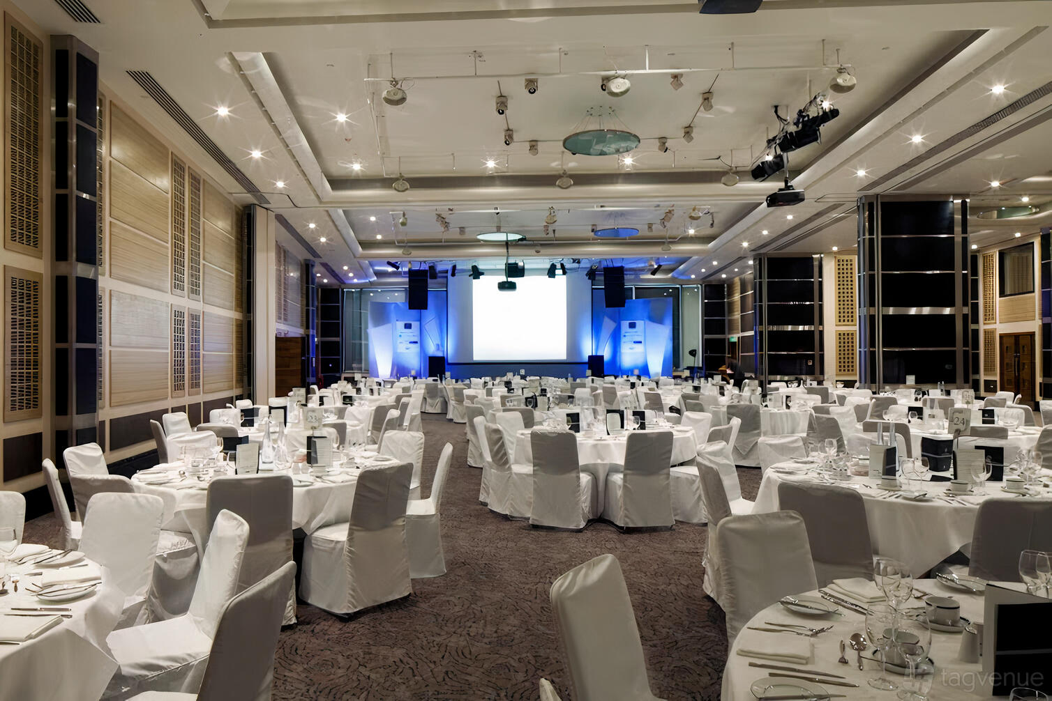 An event space with round tables draped in white linens, stage, and ceiling spotlights at Leonardo Royal London St Paul's.