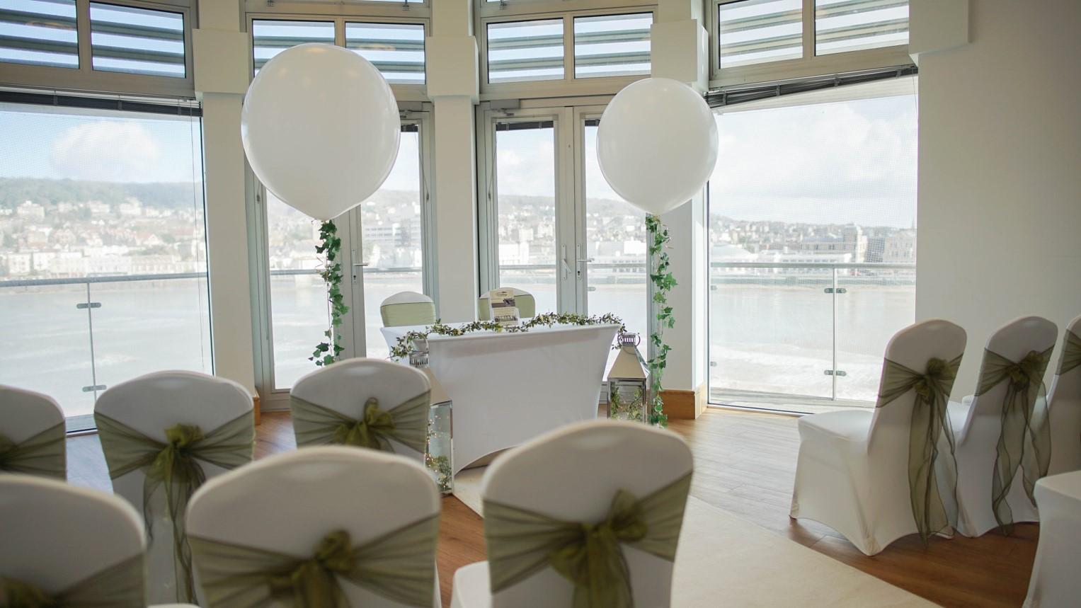 A boardroom with floor-to-ceiling windows, white chairs with green sashes, and large white balloons at The Grand Pier.