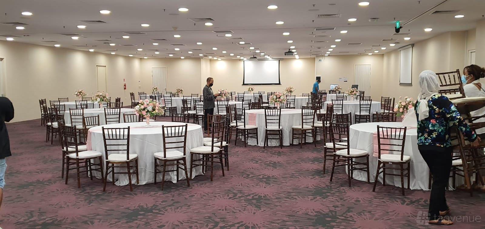A spacious hall with round tables covered in white linens, floral centerpieces, and a projector screen at THE LEGACY, One-North.