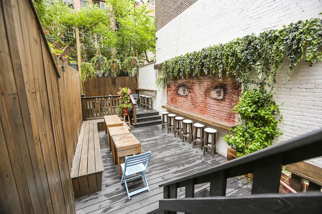An outdoor terrace with wooden seating, bar stools, and a mural of eyes on a brick wall at Hidden Lane Bar.