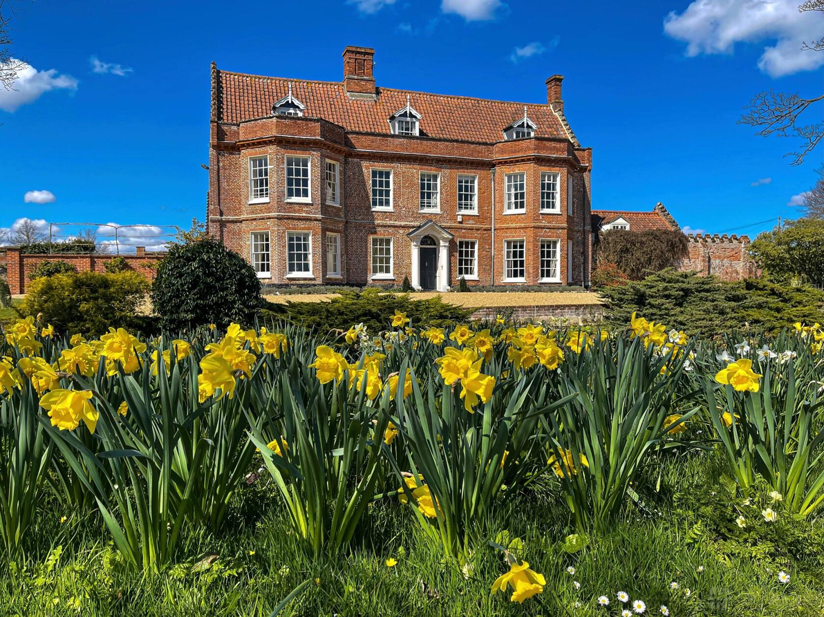 A historic red-brick barn with a tiled roof and manicured garden with blooming yellow daffodils at Elms Barn.