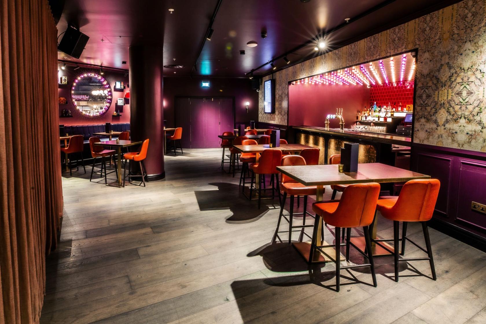 An event venue with purple walls, patterned wallpaper, high tables with orange chairs, and a lit bar at Puttshack Bank.