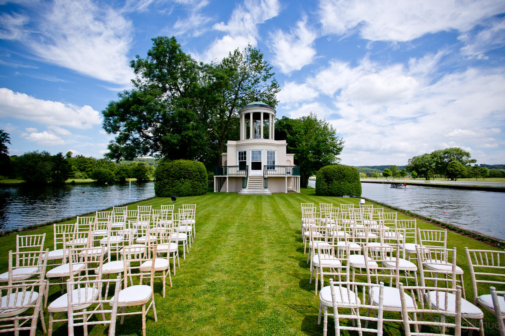 A riverside event lawn with rows of white chairs facing a pavilion surrounded by trees at Temple Island.