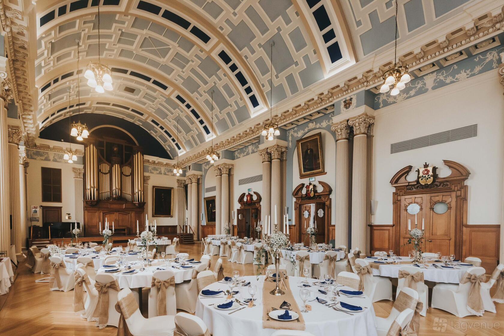 A grand hall with arched ceilings, ornate columns, and round tables dressed in white linens at Colchester Weddings.