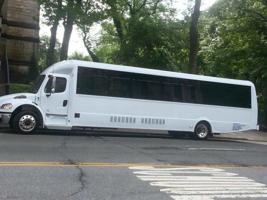 A white party bus with tinted windows parked on a city street under tall trees at US Bargain Limo.