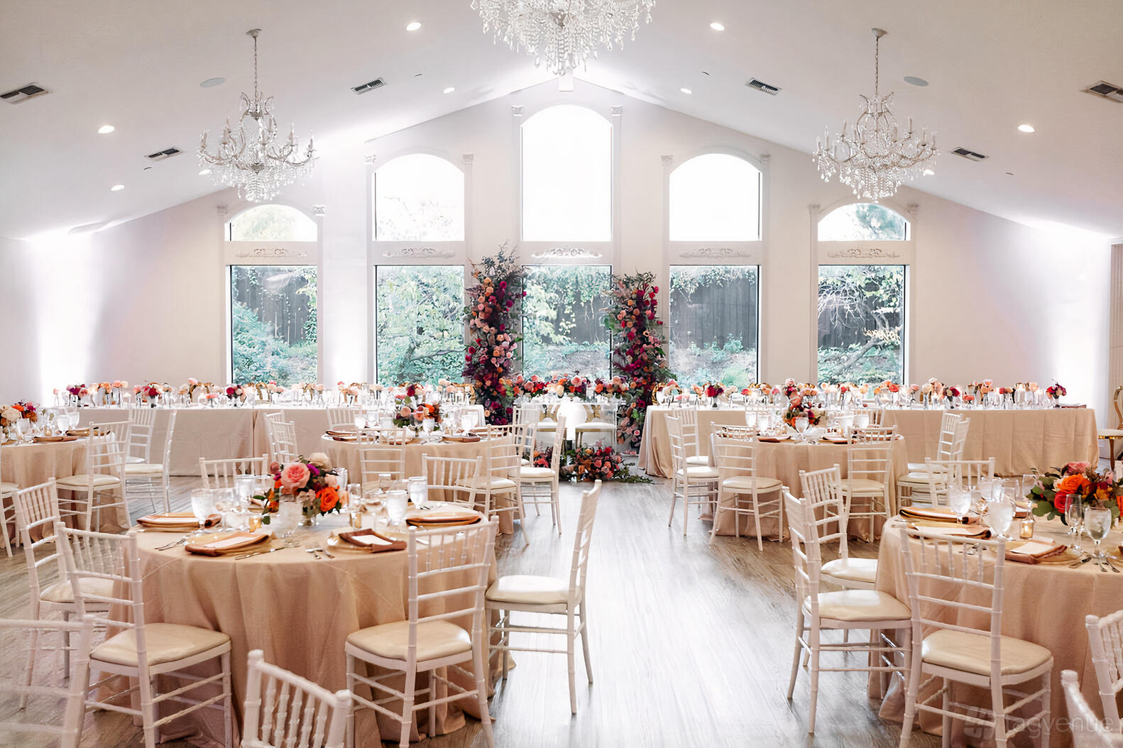 An event venue with white chiavari chairs, round tables with blush linens, and a floral arch by tall windows at Brighton Abbey.