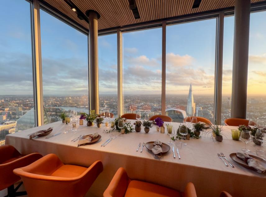 A dining room with floor-to-ceiling windows, panoramic city views, and a table set with plants at 8 Bishopsgate.