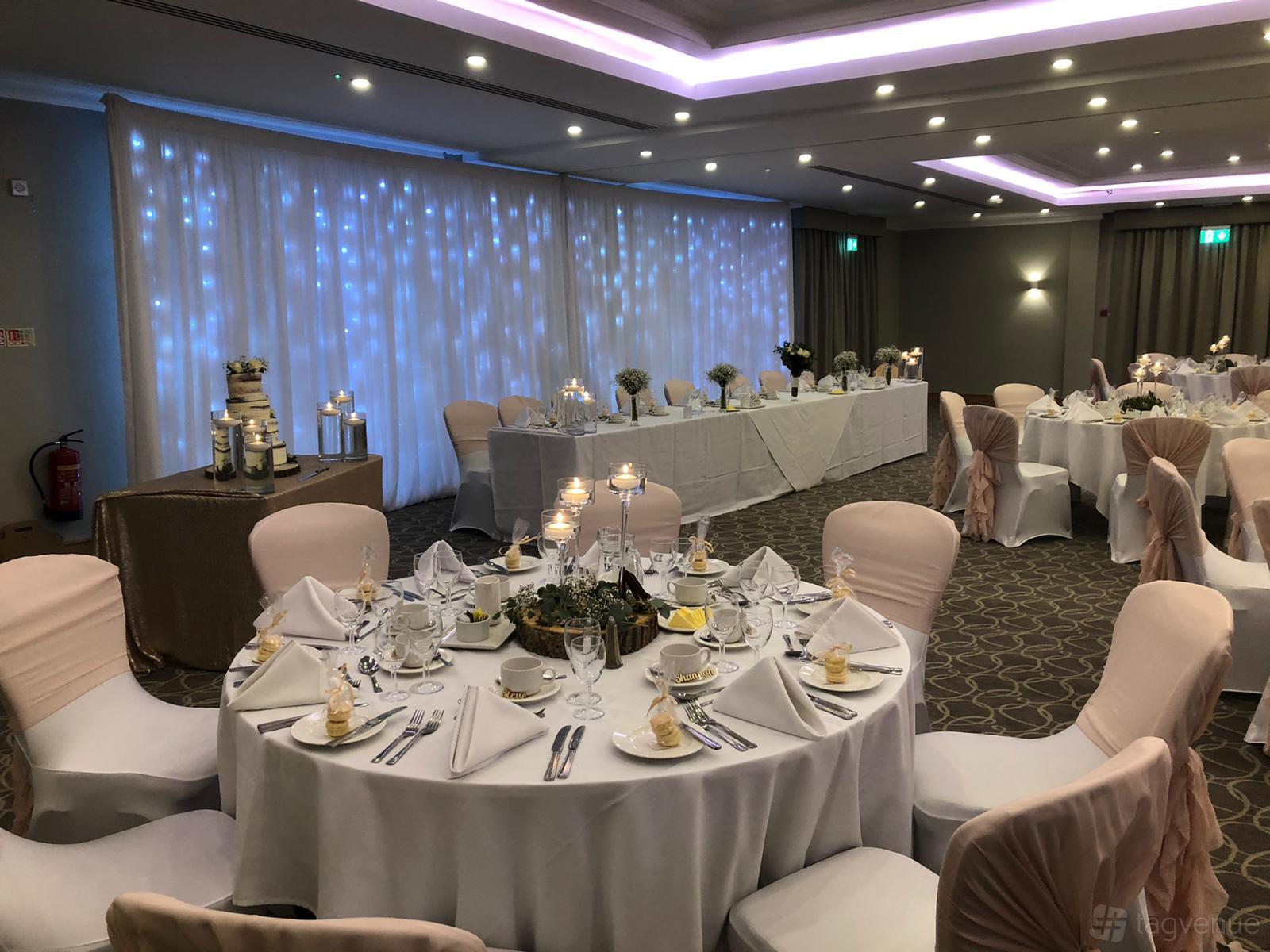 An event space with round tables dressed in white linens, chair covers, and a backdrop of twinkling lights at Hellidon Lakes Golf & Spa Hotel.