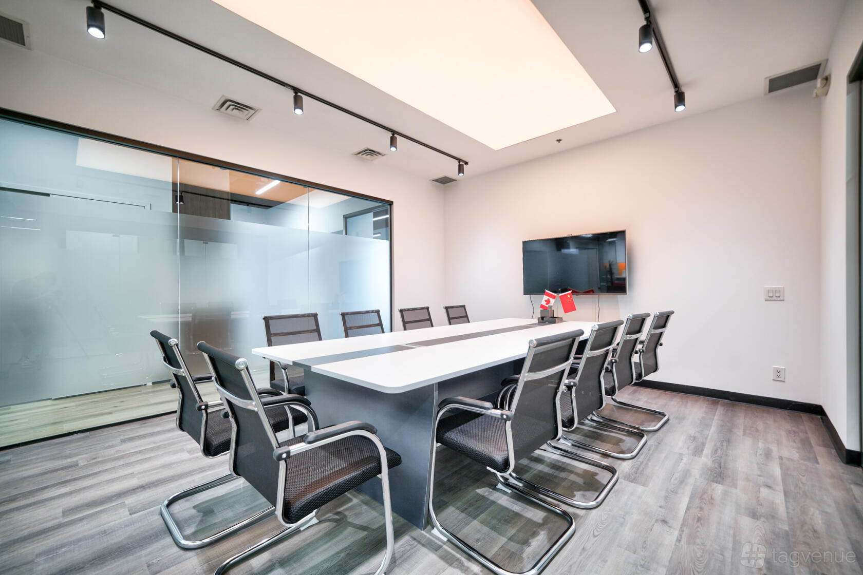 A boardroom with a long white conference table, mesh chairs, glass walls, and a wall-mounted TV at LaunchHub Space.