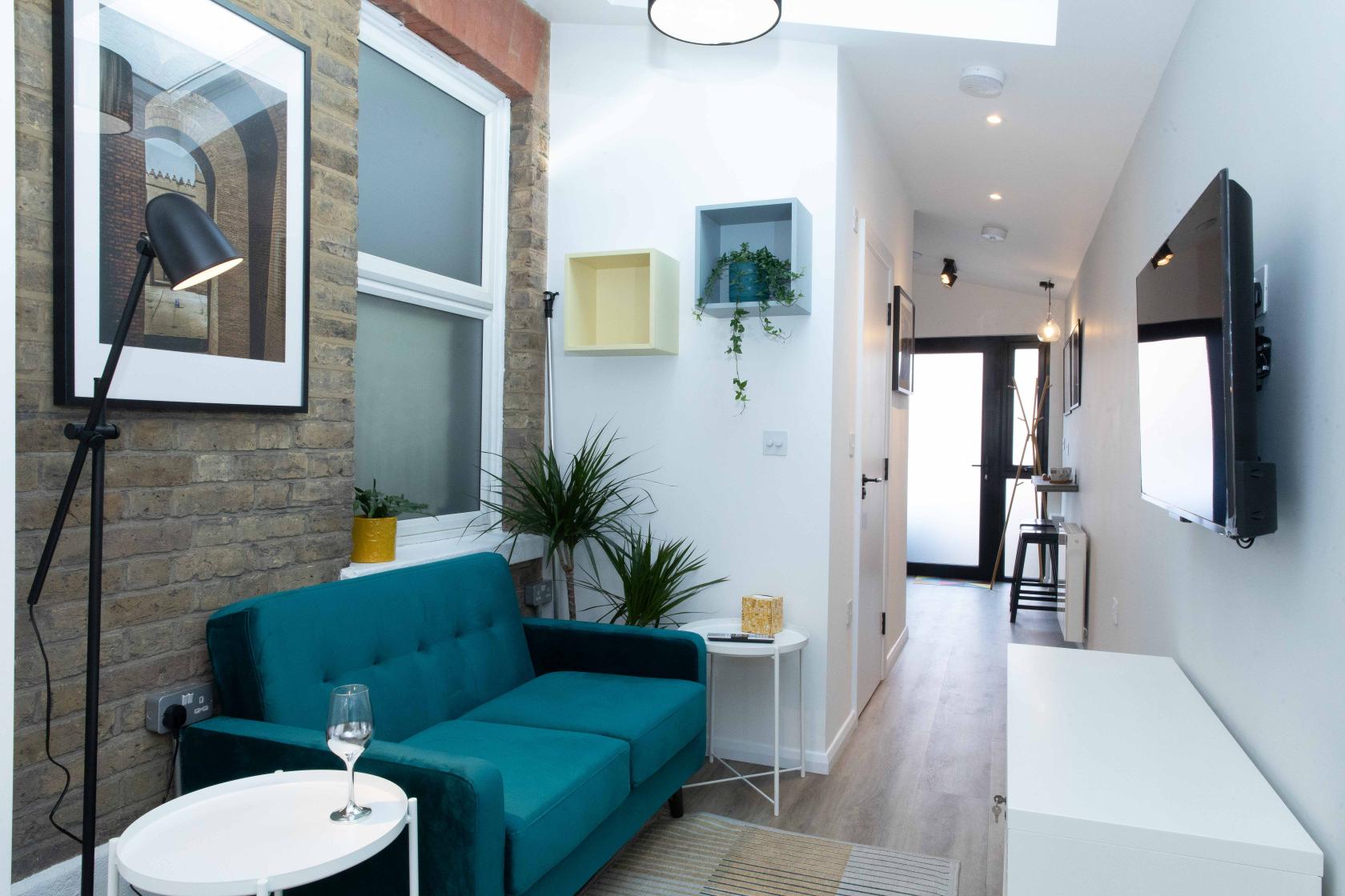 An apartment living area with exposed brick wall, teal sofa, potted plants, and wall-mounted TV at The Annex.