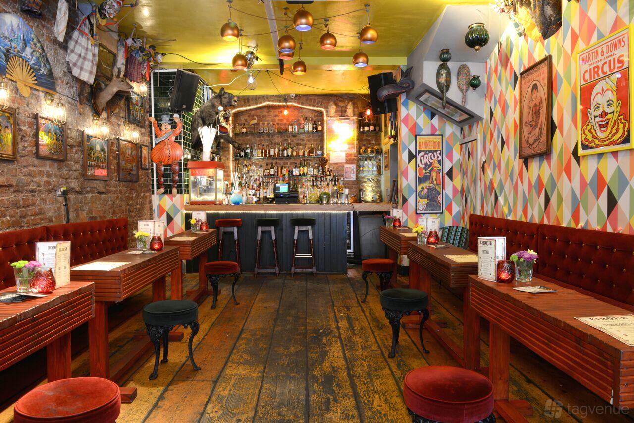A cocktail bar with exposed brick walls, patterned wallpaper, vintage circus decor, and wooden tables at Cirque.