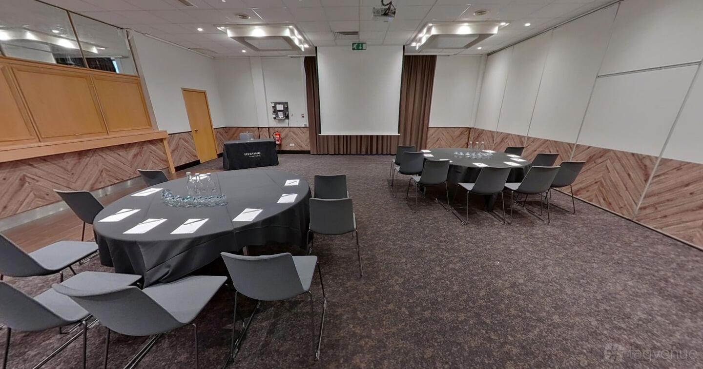 A meeting room with round and rectangular tables, grey chairs, and a projector screen at Novotel Birmingham Centre.