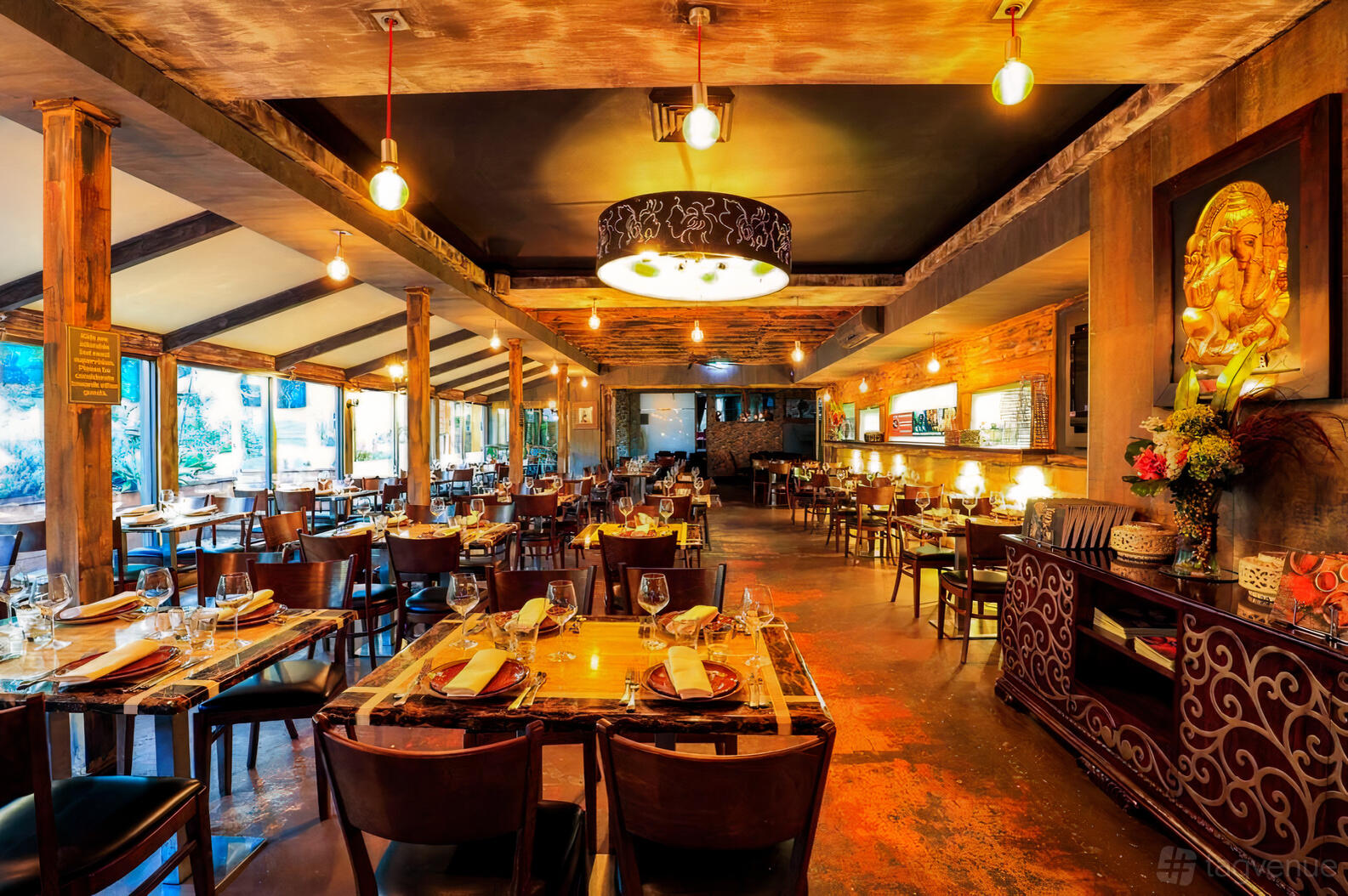 A dining room with wooden beams, exposed brick walls, warm lighting, and neatly set tables at Urban Tadka.