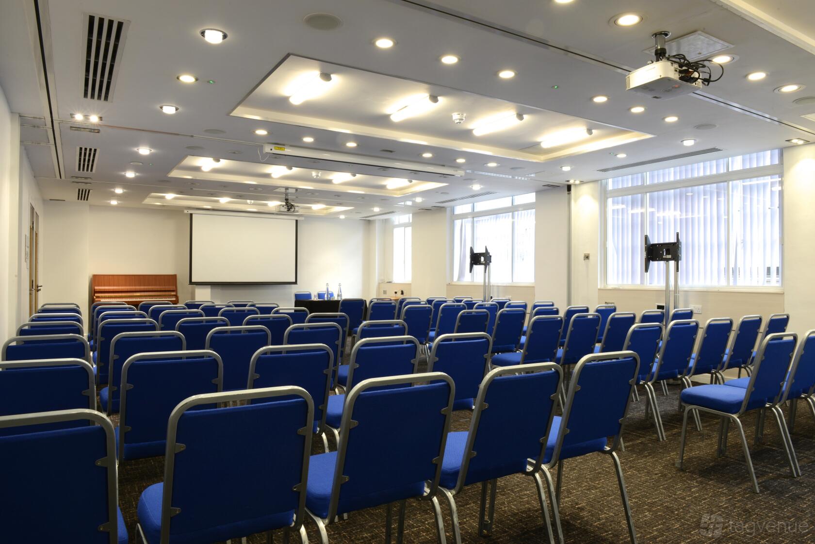 An event space with rows of blue chairs, a projector, and large windows at The Wesley Euston Hotel.