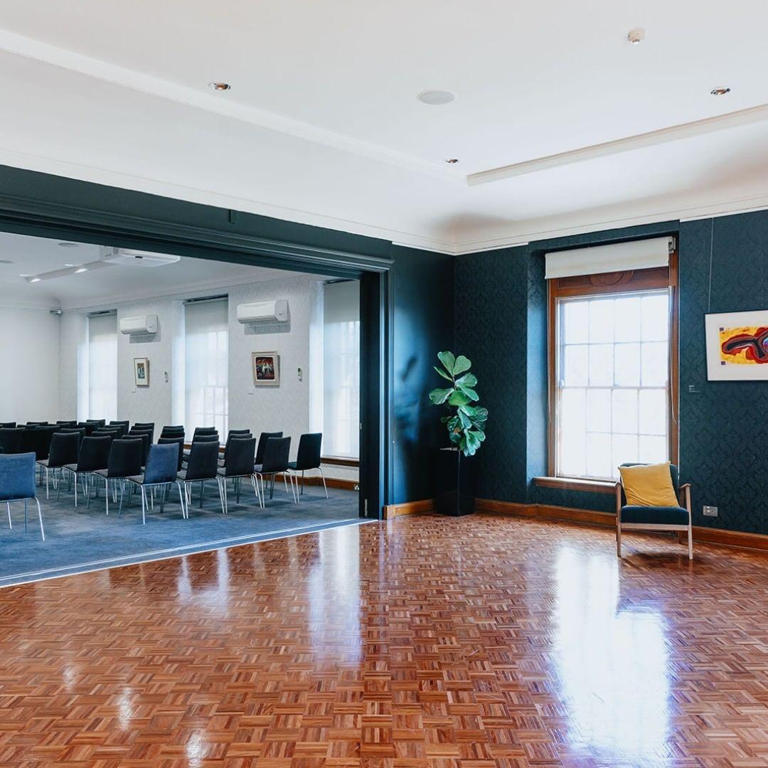 A meeting room with parquet flooring, rows of black chairs, large windows, and framed artwork at HostCo Sydney.