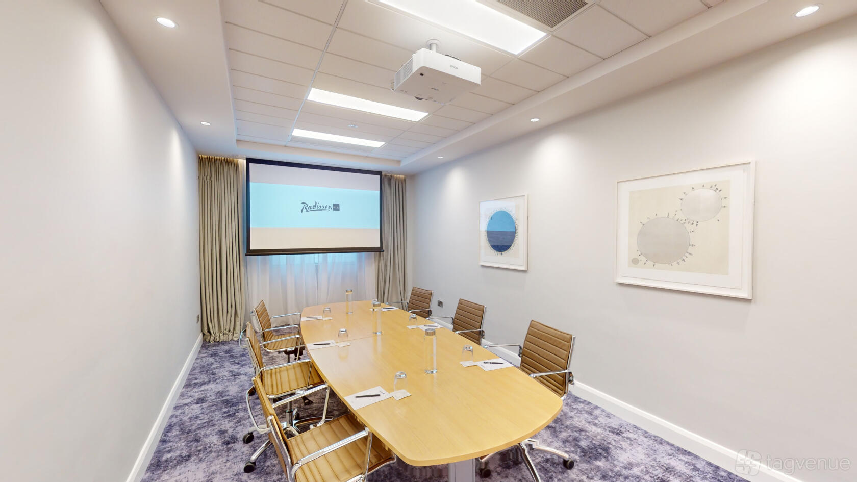 A meeting room with an oval conference table, ergonomic chairs, a ceiling projector, and wall art at Radisson Blu Hotel, Dublin Airport.