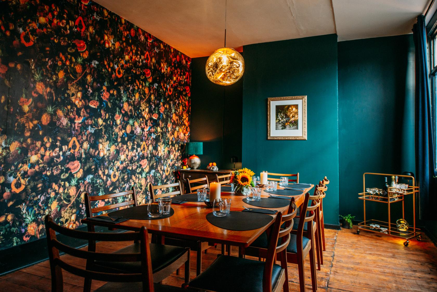 A dining room with a floral accent wall, large wooden table, and gold pendant light at Gulp.