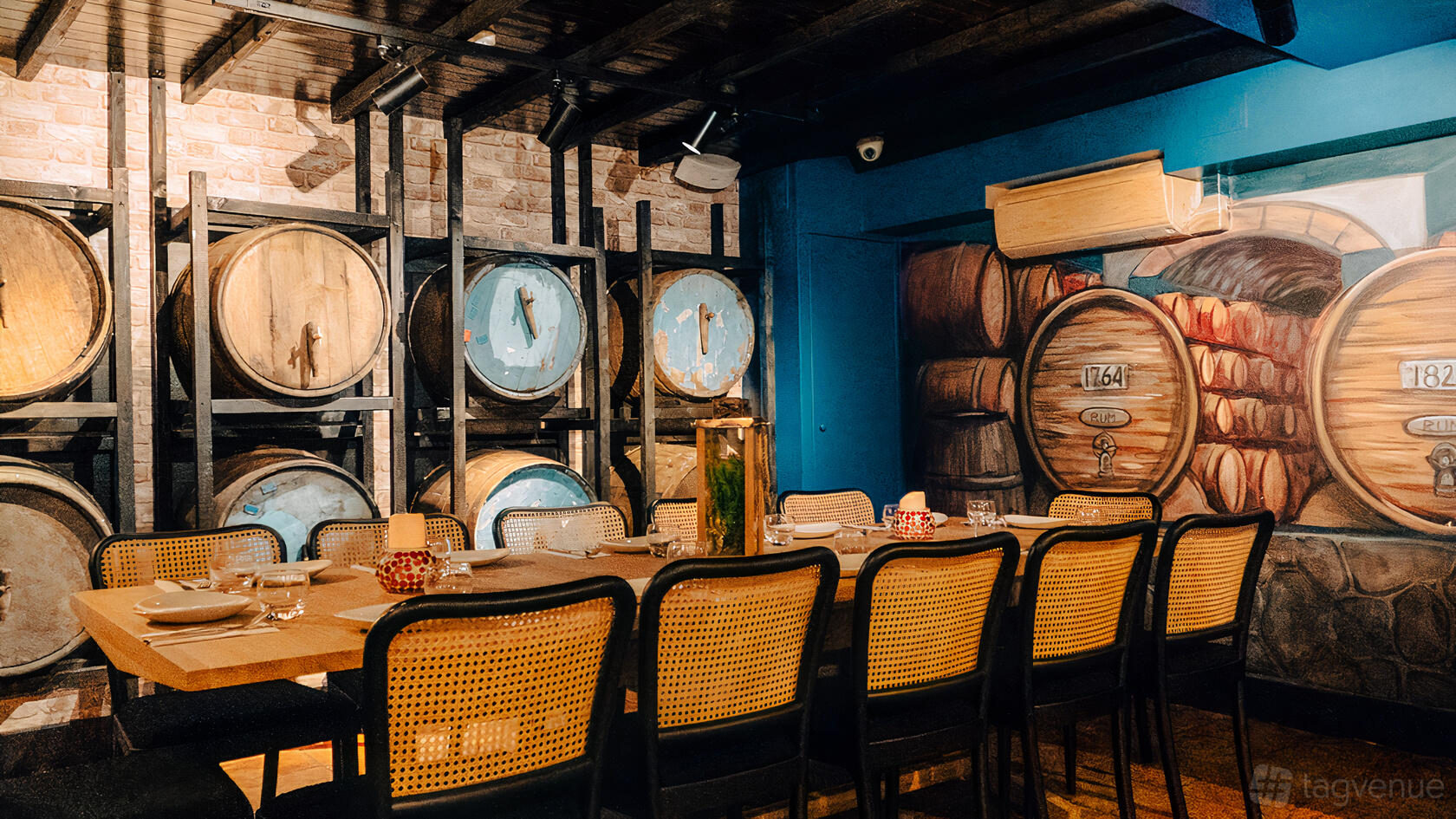 A restaurant dining room with a long wooden table, rattan chairs, barrel wall decor, and exposed brick at Cottons Notting Hill.