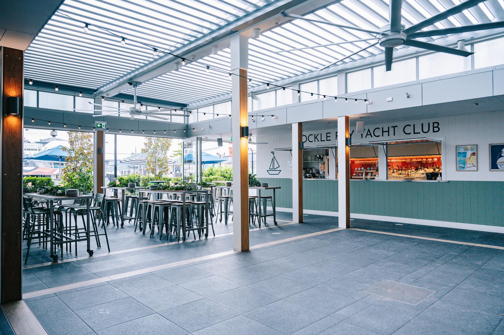 An open-air bar with high tables, stools, string lights, and a covered roof at Cockle Bay Yacht Club.