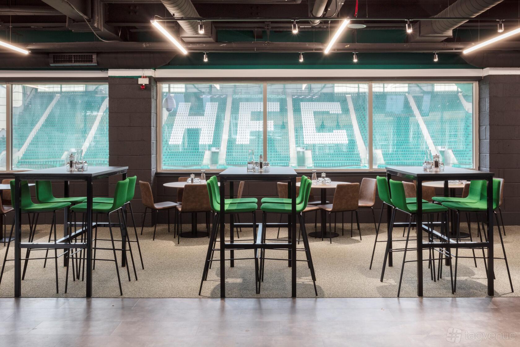 A stadium event lounge with high tables, green chairs, and large windows overlooking the stands at Easter Road Stadium.