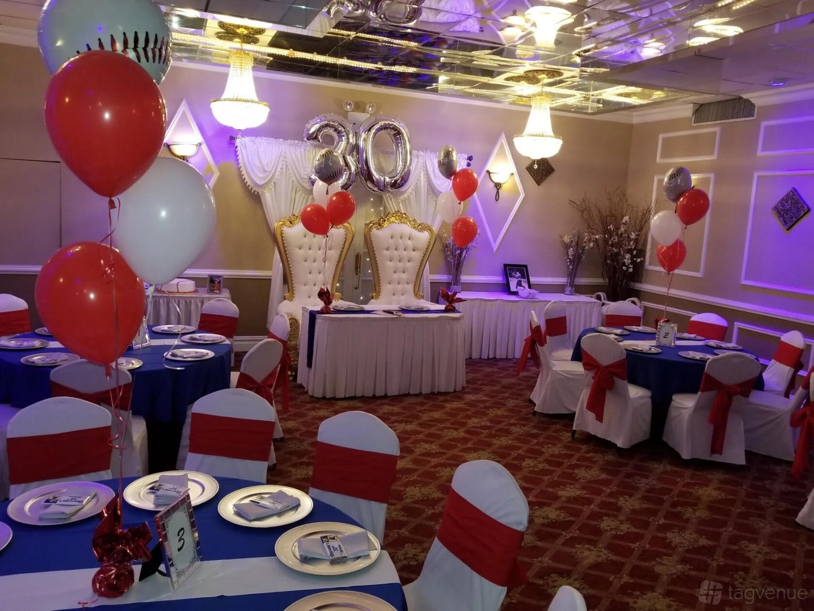 A banquet hall with balloon decorations, white and red chair covers, and two ornate chairs at The Grand 2020 Banquet & Party Hall.
