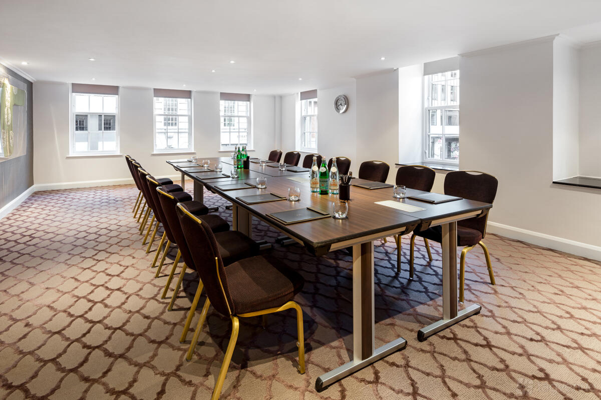 A meeting room with large windows, patterned carpet, and a long conference table at The May Fair Hotel.