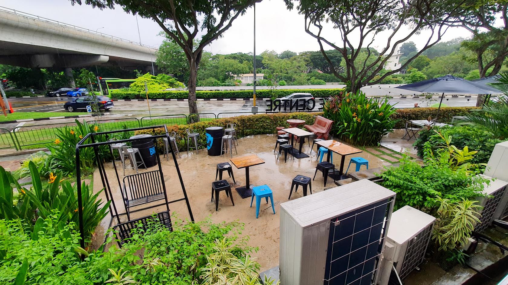 An outdoor garden patio with mixed tables and stools, greenery, and street views at Orh Gao Taproom Craft Beer Bar.