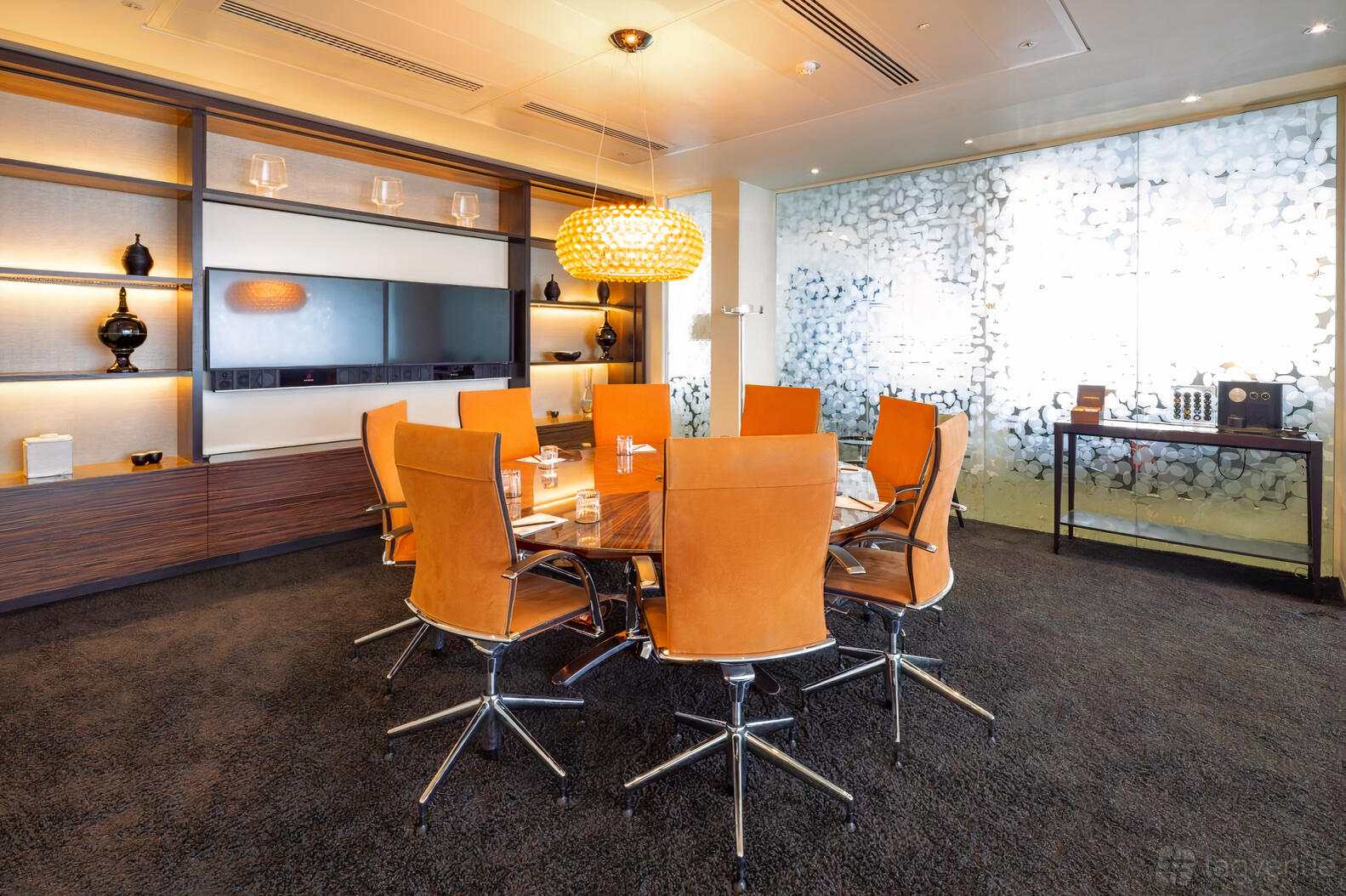 A meeting room with a round wooden table, leather chairs, built-in shelves, and a glass wall at Landmark: Heron Tower, London.
