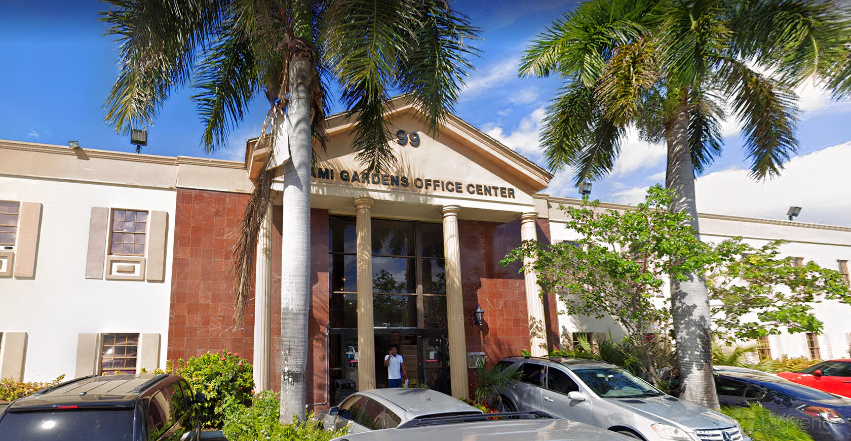 An office building entrance with columns, glass doors, palm trees, and a sign reading Miami Gardens Office Center at We Major Studios