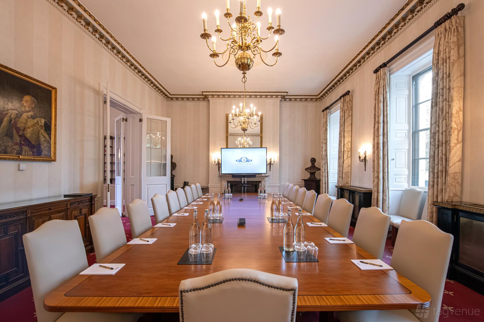 A meeting room with a long wooden boardroom table, cream chairs, chandeliers, and a large screen at No.11 Cavendish Square.