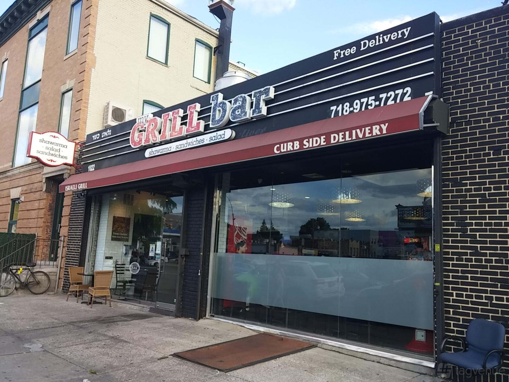 A restaurant with a glass storefront, outdoor seating, and red curbside delivery awning at My Grill Bar.