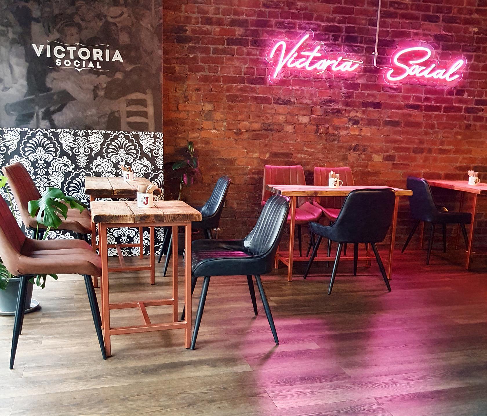 An event space in a restaurant with exposed brick walls, neon signage, and wooden tables at Victoria Social.
