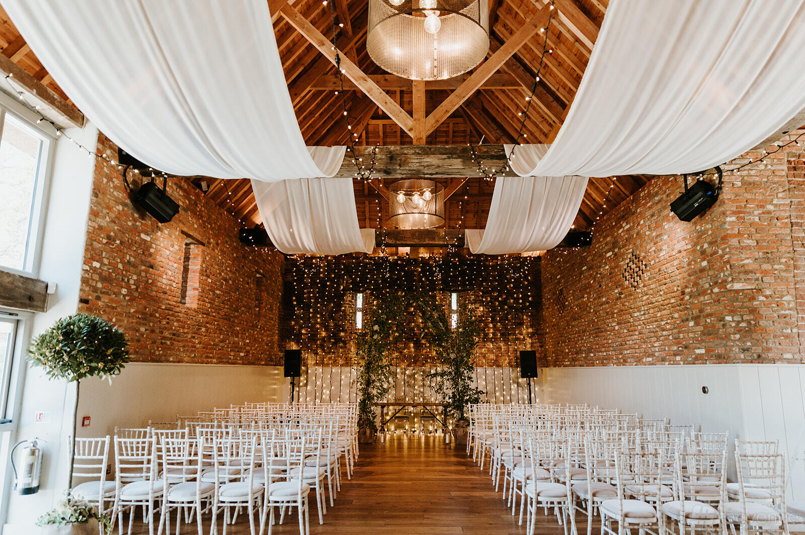 A barn venue with exposed brick walls, draped white fabric ceiling, wooden beams, and rows of white chairs at Worstead Estate.