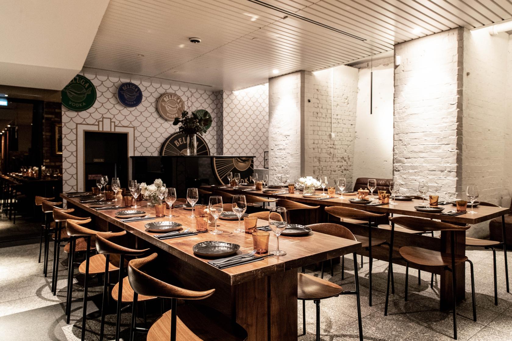 A dining room with long wooden tables, modern chairs, exposed white brick walls, and floral centerpieces at The George on Collins.