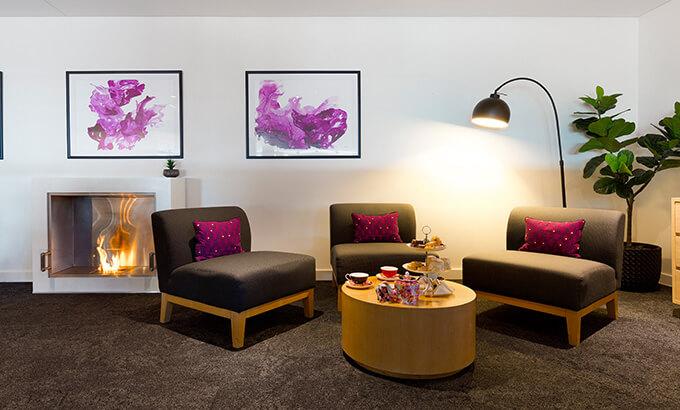 A meeting room with a cozy lounge area, modern fireplace, round table with refreshments, and wall art at Sage Hotel Wollongong.
