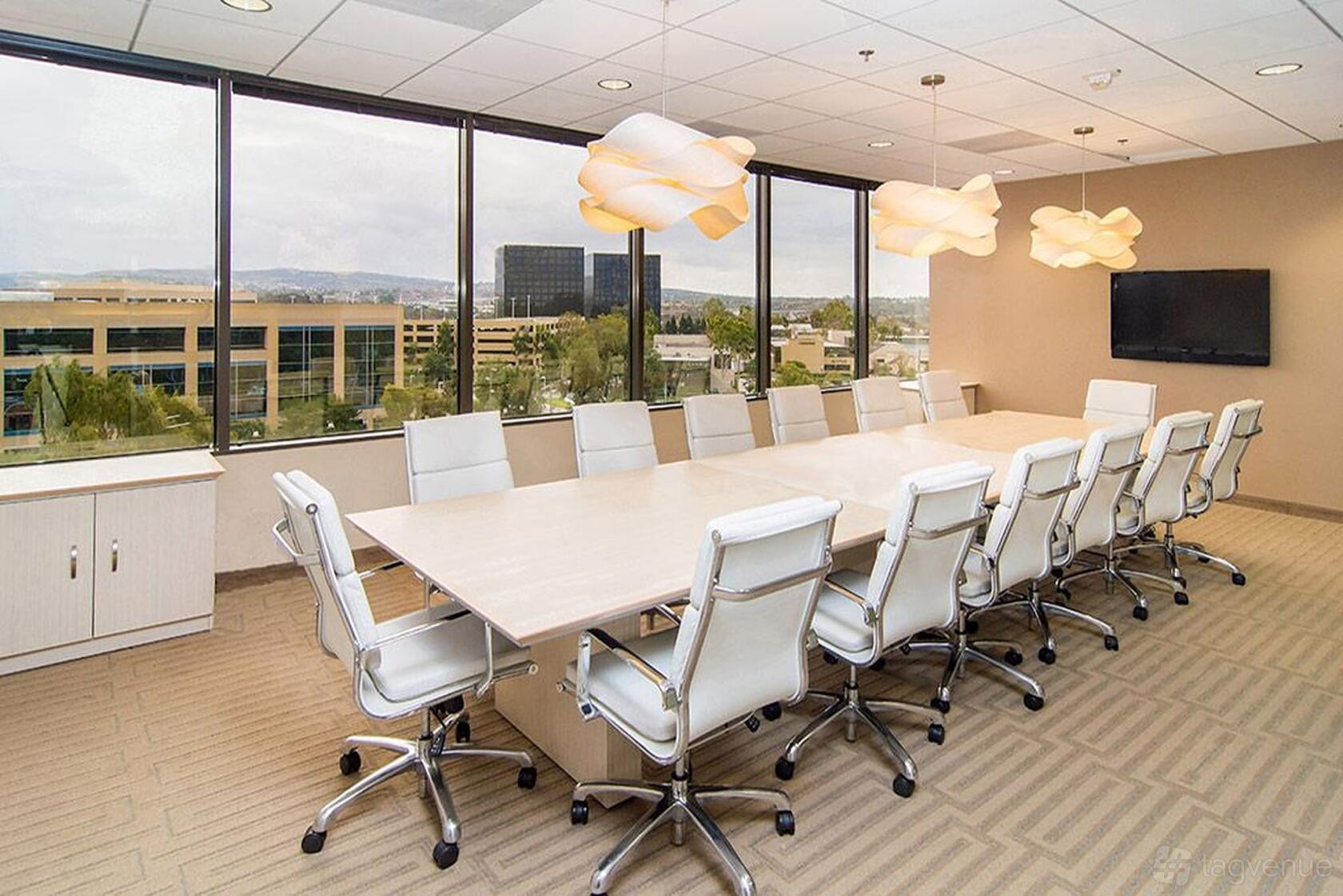 A coworking space with a large conference table, ergonomic chairs, modern pendant lights, and wall-mounted TV at Premier Workspaces - 4000 MacArthur Boulevard.