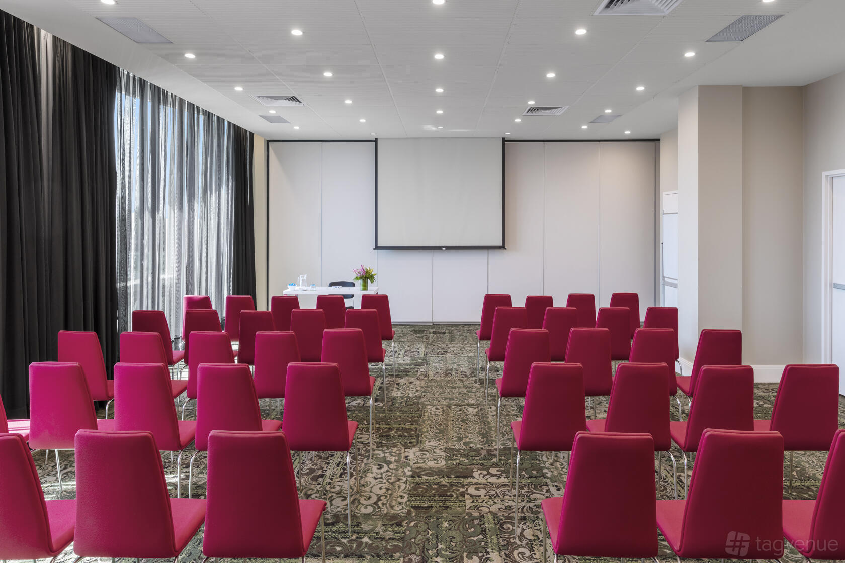 A function room with rows of red chairs, patterned carpet, and a projector screen at Nesuto Parramatta Apartment Hotel.