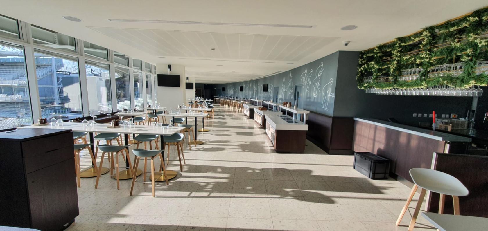 An event space with floor-to-ceiling windows, high tables and stools, and cricket-themed wall art at Lord's Cricket Ground.