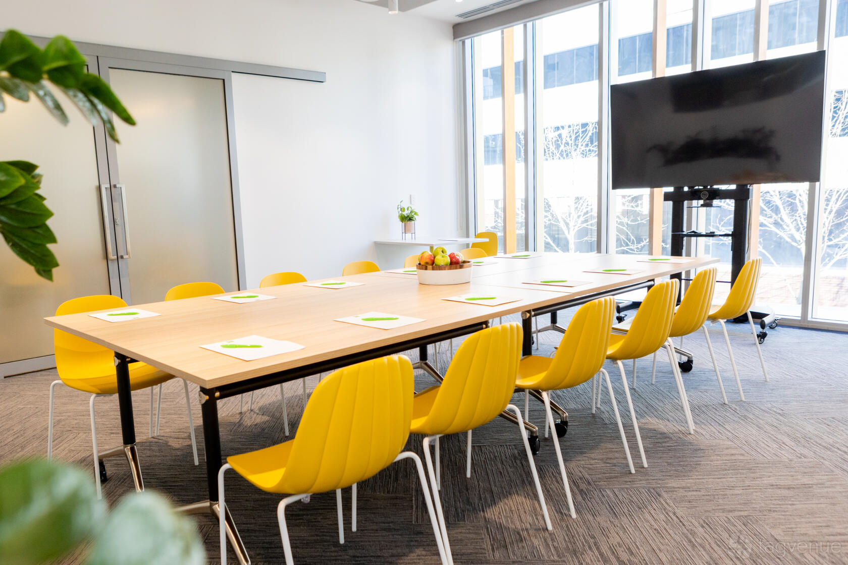 A meeting room with yellow chairs, a rectangular table, floor-to-ceiling windows, and a large TV screen at ibis Styles East Perth.