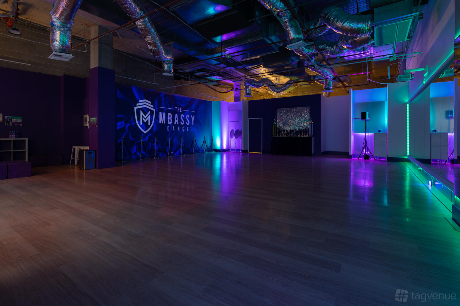 A dance studio with wood floors, colorful LED lighting, mirrored walls, and exposed ceiling ducts at The MBassy Dance.